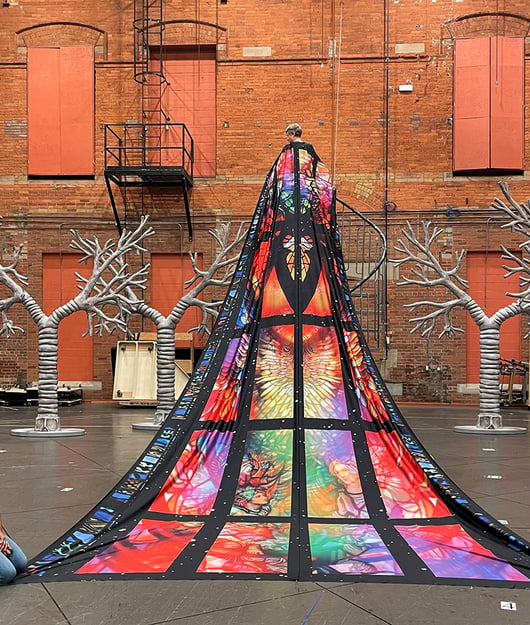 An actor on the stage wears a long sweeping cloak that is designed like a stained glass window.